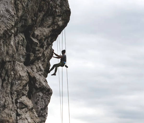 picture of a person climbing hanging from ropes on his harness, resting in midair facing a rocky mountain